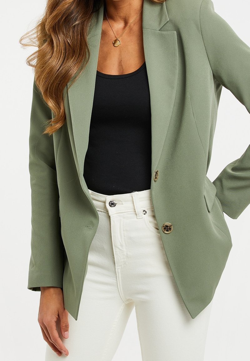 Blazer vert olive avec un design ajusté, simple boutonnage, deux boutons dorés, associé à un haut noir et un pantalon blanc.