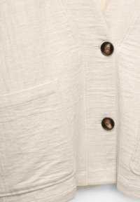 Lichtbeige button-up jasje gemaakt van textuurstof, met een V-hals, grote voorkruiportefeuille en twee bruine knopen.
