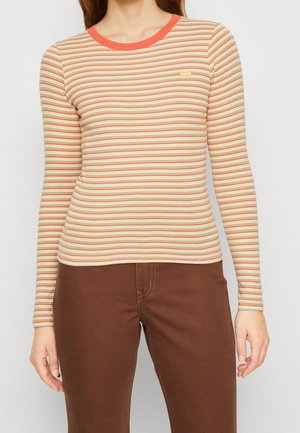 Femme portant une chemise crème ajustée à manches longues avec des rayures horizontales orange, jaune et verte, et un pantalon marron.