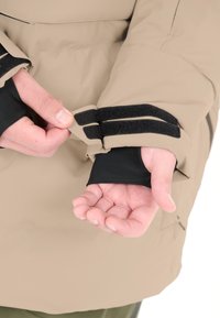 Beige ytterskal med justerbara kardborrearmar. Svart inre ärm synlig. Slät textur, minimalistisk design och praktisk stängningsdetalj.