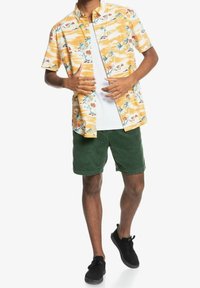 Kortärmad, gul tropisk skjorta med palm- och blommönster, tillsammans med gröna kordbyxor och svarta sneakers. Vit undertröja synlig.