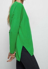 Green sweater with long sleeves, featuring a relaxed fit and side slits, made from a soft knit fabric. Worn with dark pants.