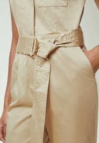 Combinaison beige avec large ceinture en tissu cousue nouée en nœud, main dans la poche, gros plan sur la taille et le torse.