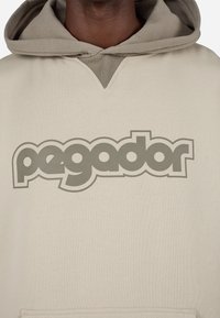 Μπέζ φούτερ με μεγάλο γκρίζο λογότυπο "pegador" μπροστά. Ο σχεδιασμός διαθέτει άνοιγμα σε σχήμα V και μαλακό υφασμάτινο υλικό.