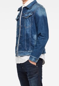 Man med en blå denimjacka över en vit skjorta och mörkblå jeans, med handen i fickan, mot en enkel vit bakgrund.