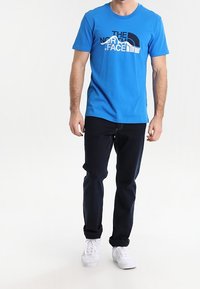 Blå bomulls-T-shirt med grafisk logotyp, kombinerad med mörka jeans och vita sneakers. Avslappnad passform med korta ärmar.