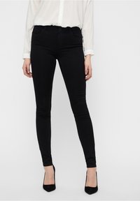 Jeans skinny en denim noir avec une texture lisse, dotés d'une taille mi-haute, de cinq poches et d'une fermeture classique par bouton et zip.