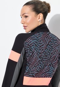 Donna con i capelli raccolti a chignon che indossa una giacca nera con strisce grigie e corallo, e un motivo astratto ondulato, mentre guarda di lato su uno sfondo bianco.
