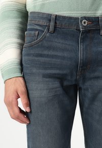 Jeans in denim blu scuro con un classico design a cinque tasche, caratterizzati da cuciture arancioni e una superficie liscia. Mano appoggiata sulla coscia.