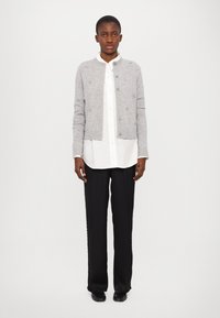 Grå strikket cardigan med teksturerede detaljer, knaplukning, ribkantede manchetter, båret over en hvid skjorte og sammen med sorte, bredbenede bukser.