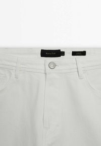 Jean blanc slim avec fermeture par boutons à l'avant et passants pour ceinture, montrant les étiquettes de marque et de coupe à l'intérieur de la ceinture.