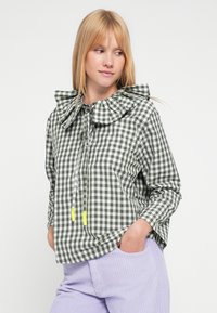 Camisa de cuadros vichy verde y blanca con un gran cuello fruncido, cordón ajustable y tancas amarillas contrastantes. Corte holgado, mangas largas.