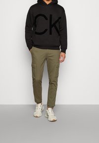 Sudadera negra con capucha y un logo gigante de CK, pantalones cargo verde oliva con bolsillos laterales y zapatillas blancas con detalles en varios colores.