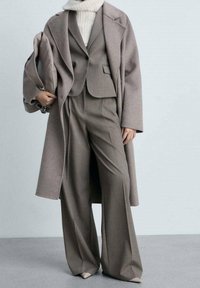 Ruskea villakangastakki, joka on puettu yhteensopivan räätälöidyn puvun päälle, jossa on leveälahkeiset housut. Ylin kerros on ylipitkä, ja siinä on kaulus sekä rento istuvuus.