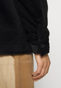 Sort fløjlsjakke med ribbet tekstur, knapmanchetter og en afslappet pasform, parret med beige bukser med kontrasterende syninger.