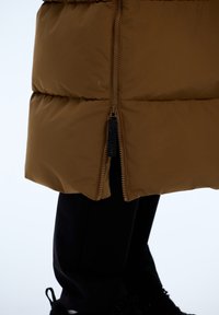 Bruine pufferjas met een verticale ritssluiting, voorzien van gequiltte secties. Gecombineerd met zwarte broek en donkere schoenen. Gladde textuur.