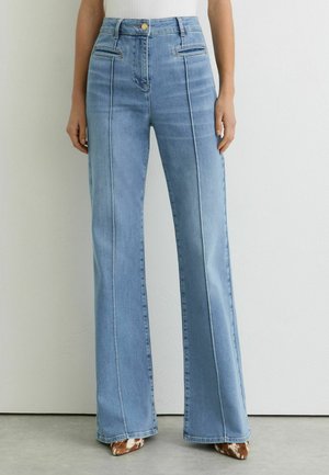Jean bleu clair taille haute à jambes larges avec des coutures sur le devant, poches avant plus petites, porté avec des chaussures pointues marron et blanches.