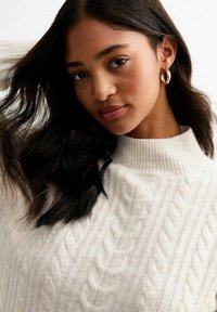 Cremefarbener Kabelstrickpullover mit hohem geripptem Kragen, der komplizierte strukturierte Muster aufweist. Das Modell trägt goldene Creolen. Dunkles, welliges Haar.