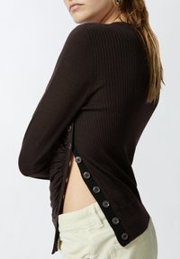 Top a coste marrone scuro con una placca di bottoni laterale con rifiniture nere e dettagli arricciati lungo il lato. Abbinato a pantaloni chiari.