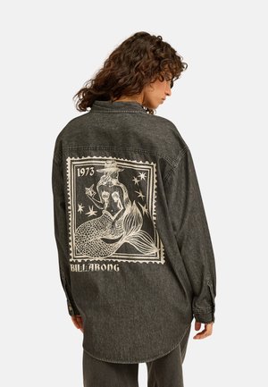Vrouw met krullend haar draagt een donker oversized spijkerjack met een grote zeemeermingrafiek en de tekst "1973 Billabong" op de achterkant.