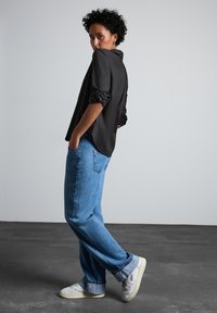 Camicia nera con colletto e maniche arrotolate, abbinata a jeans azzurri a vestibilità ampia e sneakers bianche, su uno sfondo semplice.