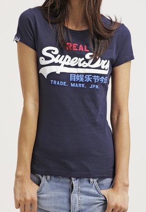 Femme portant un t-shirt SuperDry bleu marine avec un texte blanc, rouge et bleu, associé à un jean en denim bleu clair.