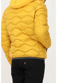 Veste matelassée jaune avec capuche et motif texturé, assortie à un pantalon noir avec un logo sur la poche arrière.