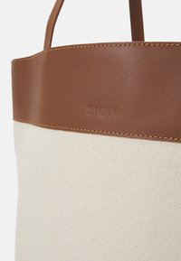 Plátěná taška s hnědým koženým akcentem a držadly. Obsahuje embosované logo "ZIGN" a kontrastní béžové šití.