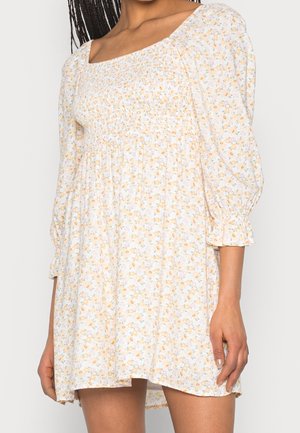 Femme portant une robe blanche à fleurs avec des manches trois-quarts bouffantes et un corsage froncé, debout devant un fond uni.