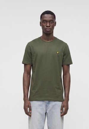 Mann steht mit Blick nach vorne, trägt ein olivgrünes T-Shirt mit kleinem gelbem Logo und hellblaue Jeans vor einem einfarbigen Hintergrund.