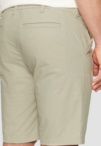 Pantalones cortos color beige claro con una tela texturizada, que presentan un bolsillo trasero, pasadores para cinturón y un corte ajustado. Detalle de costura visible a lo largo de las costuras.