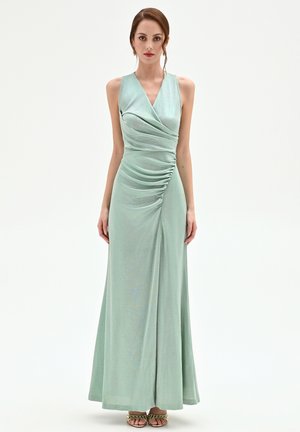 Donna che indossa un abito lungo senza maniche in raso verde pallido con dettagli plissettati sul lato destro, in piedi davanti a uno sfondo bianco uniforme.