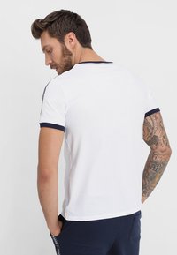 Weißes T-Shirt mit kurzen Ärmeln und marineblauen Akzenten am Kragen und an den Ärmeln, aus Baumwolle gefertigt. Die Rückseite hat ein schlichtes Design mit lockerer Passform.