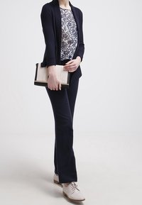 Mujer con pantalones marinos, blusa floral y cárdigan marino sujetando un bolso beige, de pie con zapatos blancos sobre un fondo claro.