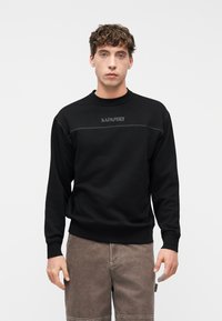 Νεαρός άνδρας με σγουρά μαλλιά, φορώντας μαύρο φούτερ Napapijri και καφέ παντελόνι, στέκεται μπροστά σε μια απλή λευκή επιφάνεια.