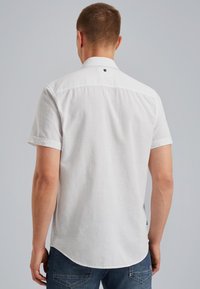 Camisa blanca de manga corta con una textura de lino ligero, con un collar estándar y un pequeño logotipo negro en la parte trasera, combinada con jeans azul oscuro.