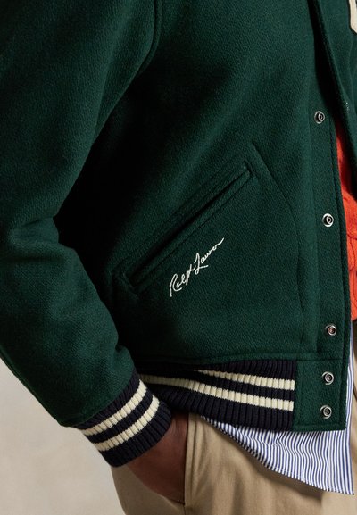 Zielona wełniana kurtka z teksturowanym wykończeniem, z białym detalem sygnaturowym, zapięciem na guziki i niebieskimi prążkowanymi mankietami.