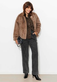 Veste en fausse fourrure marron portée sur une chemise sombre à motifs, associée à un pantalon en denim noir taille haute et des talons noirs à bout pointu. Accessoirisée avec un petit sac.