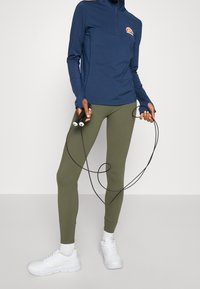 Marinblå zip-neck långärmad tröja, olivgröna leggings och vita sneakers, håller i ett hopprep. Tröjan har en broderad logotyp.