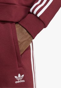 Bordeaux Adidas sportovní souprava s bílými pruhy na rukávech a po stranách. Obsahuje klokánkovou kapsu, ribkované manžety a logo na nohavici.