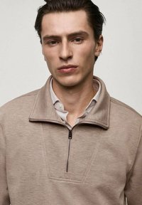 Pullover beige con colletto a metà zip, realizzato in tessuto morbido. Presenta un motivo testurizzato con un'apertura che mostra una camicia a righe leggere sottostante.