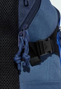 Mochila de tela azul que presenta una malla texturizada y paneles lisos. Incluye cremalleras dobles con tiradores azules y una correa de hebilla negra.