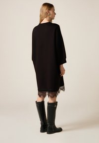 Robe noire en maille avec un col rond, ornée d'un ourlet en dentelle. Le modèle porte des bottes hautes noires, mettant en valeur le détail élégant de la robe.