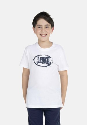 Glunderende jongen met kort donker haar, die een wit T-shirt met het logo "LEONE 1947" en donkerblauwe broek draagt, staand tegen een effen witte achtergrond.