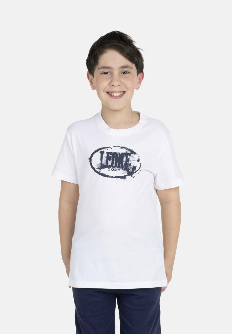 Garçon souriant aux cheveux courts et foncés portant un t-shirt blanc avec le logo « LEONE 1947 » et un pantalon bleu foncé, debout devant un fond blanc uni.