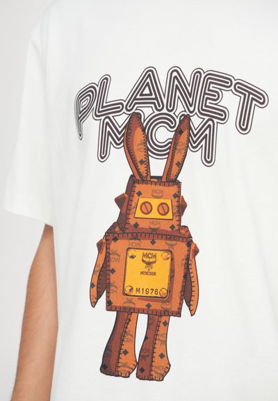 Λευκό μπλουζάκι με γραφικό σχέδιο ενός καφέ λαγού με τετράγωνο σώμα, με ετικέτες "MCM" και "München" και έντονα γράμματα που γράφουν "PLANET MCM".