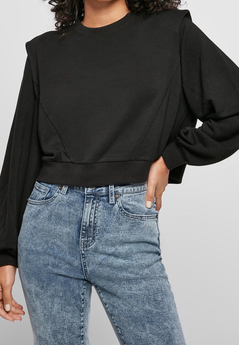 Femme portant un sweat-shirt court noir à manches longues et un jean taille haute bleu clair délavé sur un fond uni.