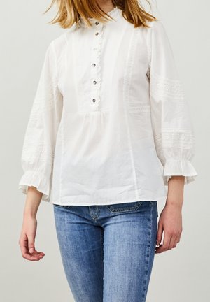 Persoon draagt een witte blouse met lange mouwen, knopen en kanten details, gecombineerd met een blauwe spijkerbroek, staand tegen een effen achtergrond.