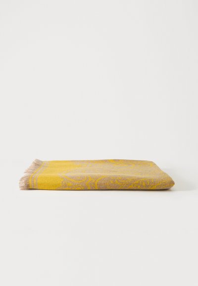 Couverture jaune et beige à franges, pliée, avec des motifs floraux complexes sur un fond blanc uni.