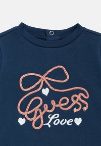 Μπλε βαμβακερό πουκάμισο με κεντημένο το κείμενο "Guess Love", σχέδιο με ροζ φιόγκο και λευκές καρδιές, στρογγυλή λαιμόκοψη και κούμπωμα με κουμπιά.